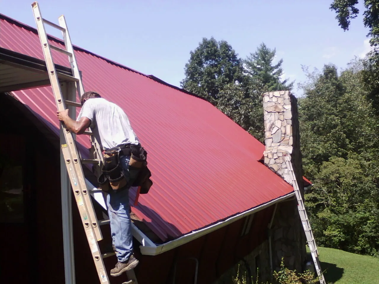 Roofer on a ladder beside a red metal roof and stone chimney — Soffit & Fascia Repair by Roofing Brothers in Langsville