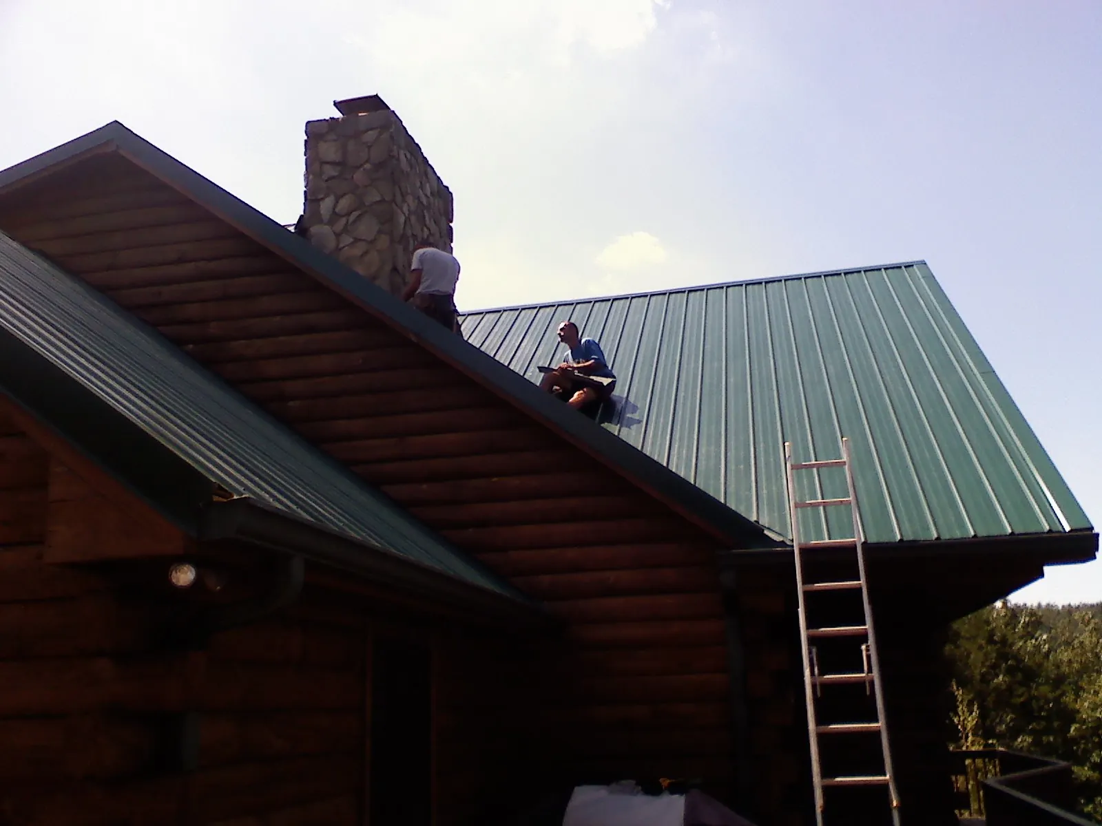 Roofers installing green metal panels beside a stone chimney on a log home in Langsville
